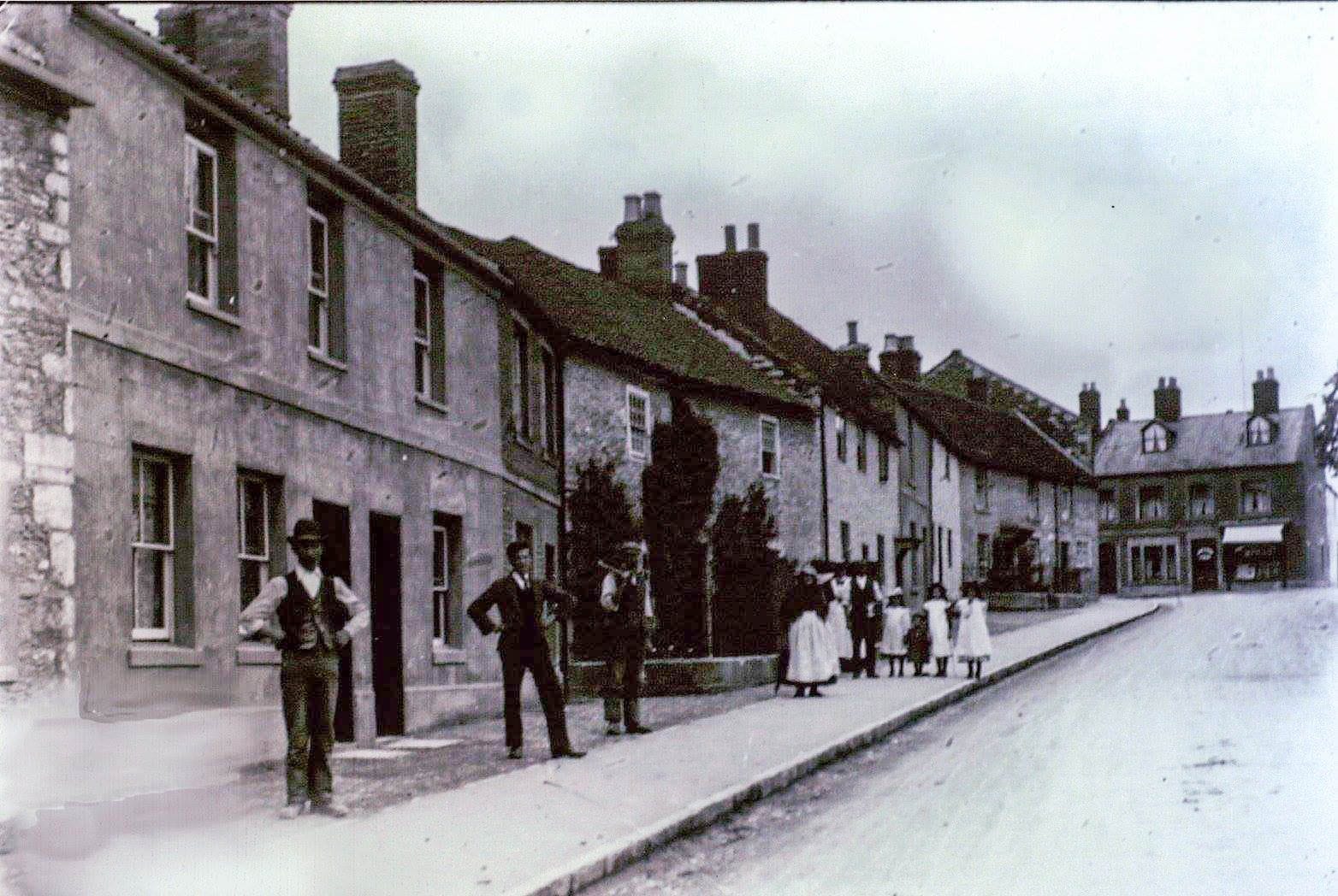 A hand-tinted view along a terrace, with men in waistcoats in the foreground and a group of women and children further along.